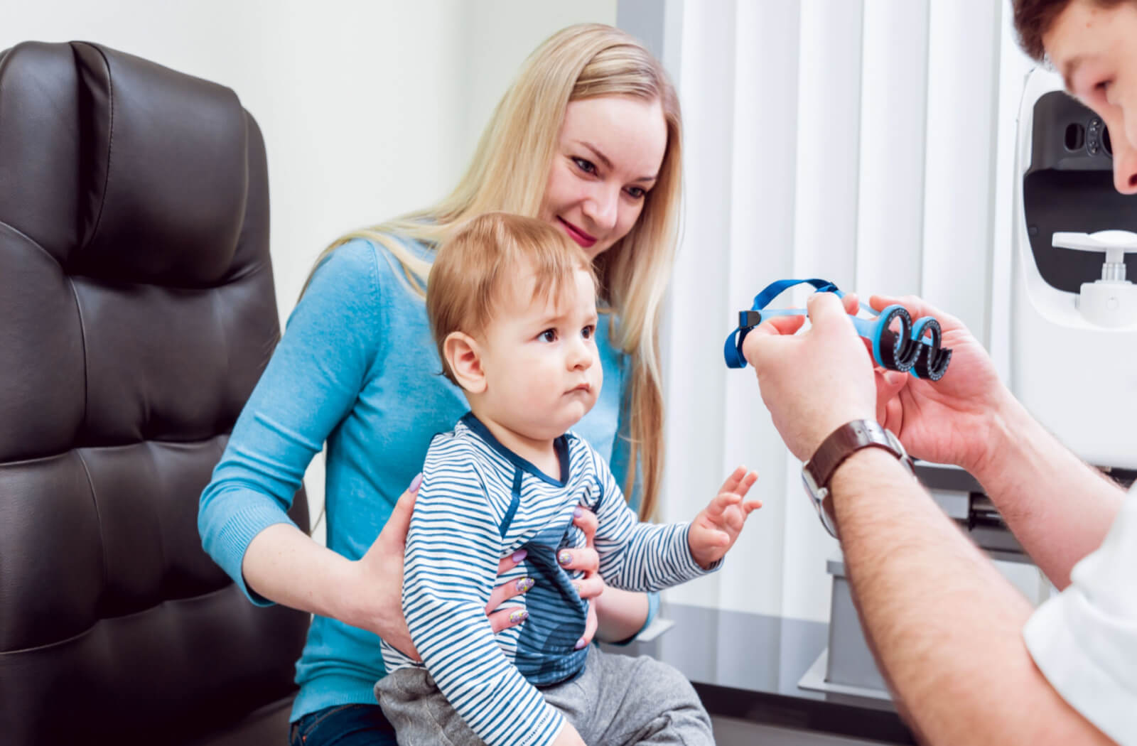 a toddler sits on his mothers lap while having an eye exam done by an optometrist