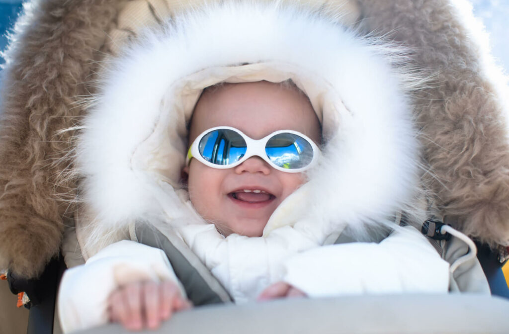 A baby in a warm stroller wearing a fur hood snow suit and a white frame infant sunglasses to protect his eye from drying.