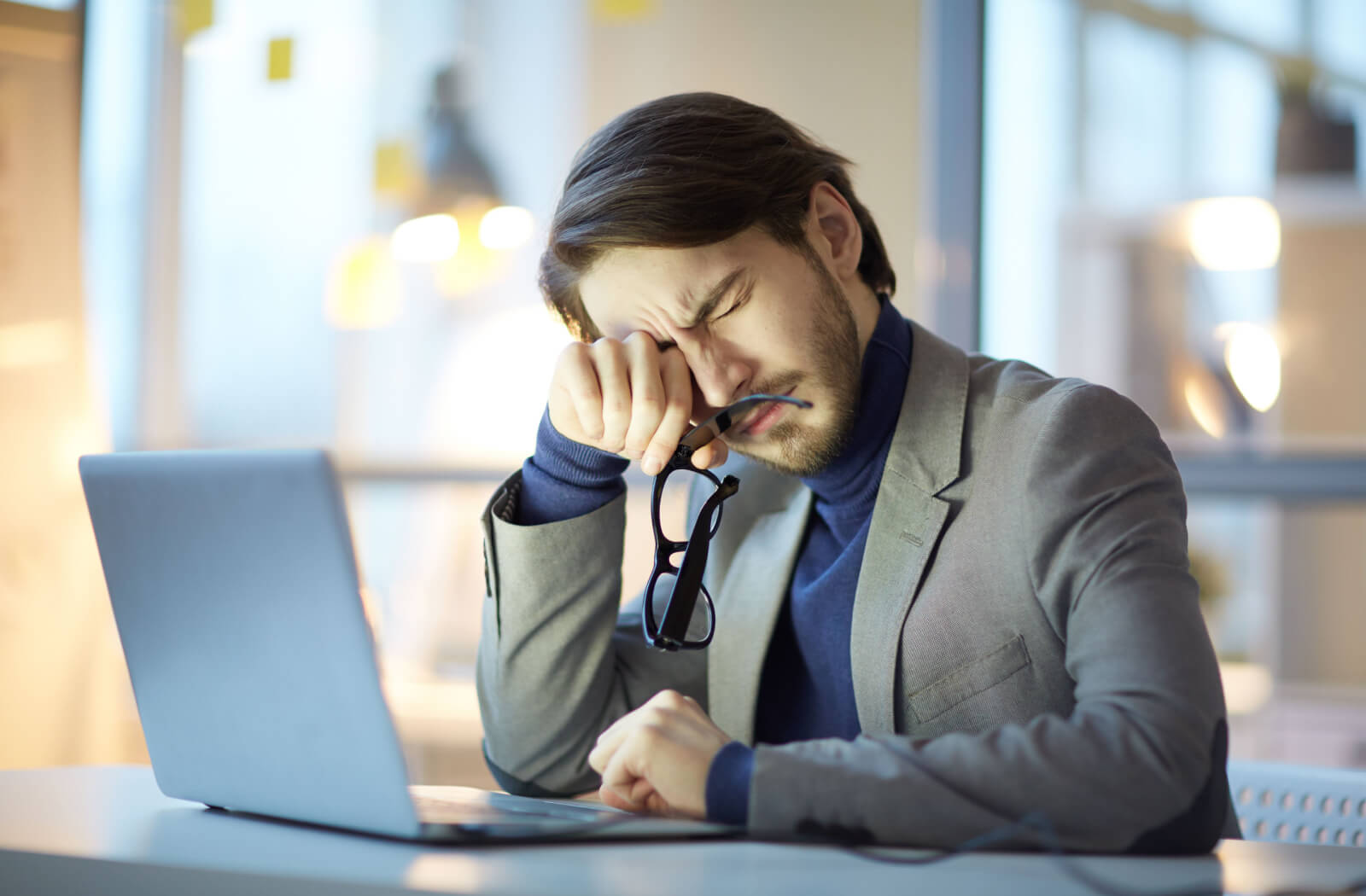 A man sitting at a desk with his laptop holding his glasses in his right hand as he rubs his eyes