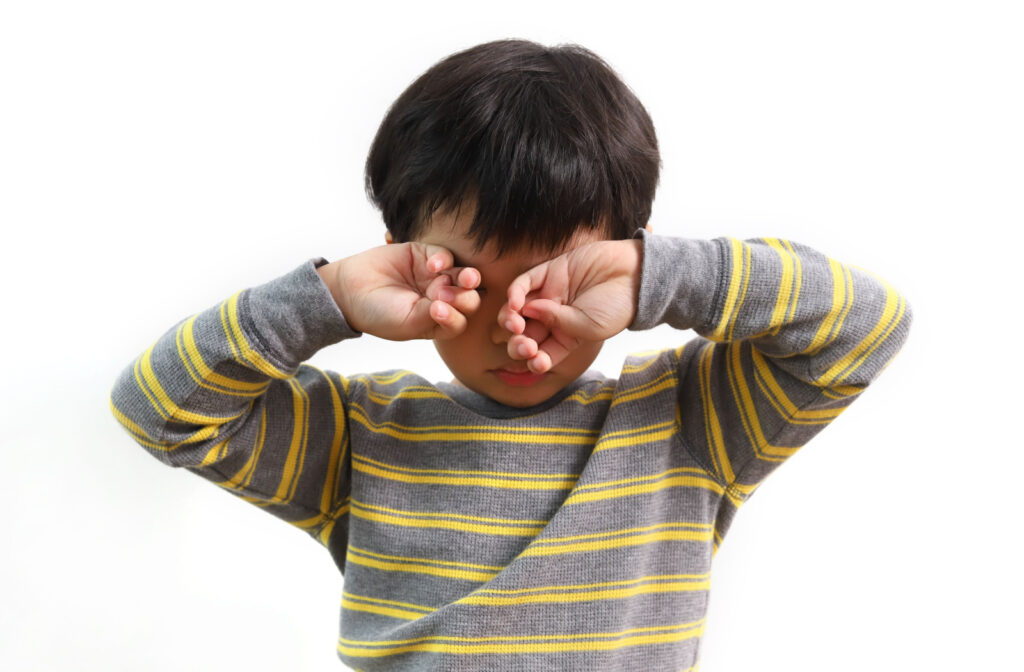 A child wearing a striped shirt holding up both arms as he rubs his eyes