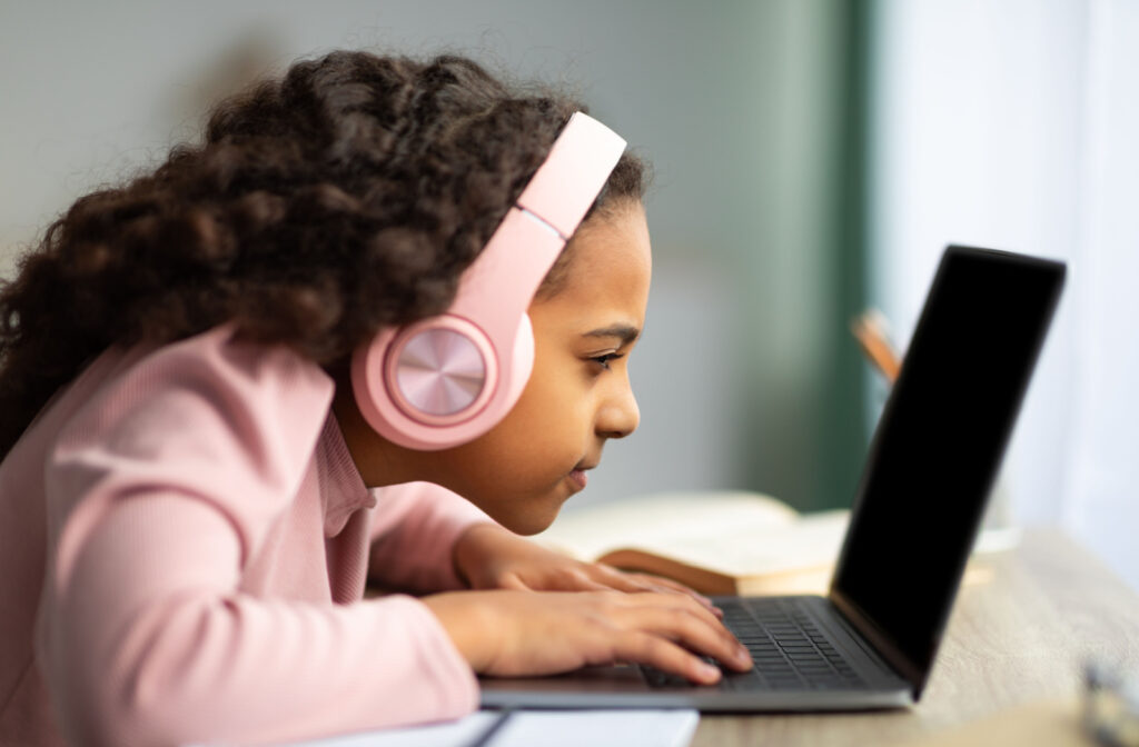 A side view of a child with headphones using a laptop very close to her face.