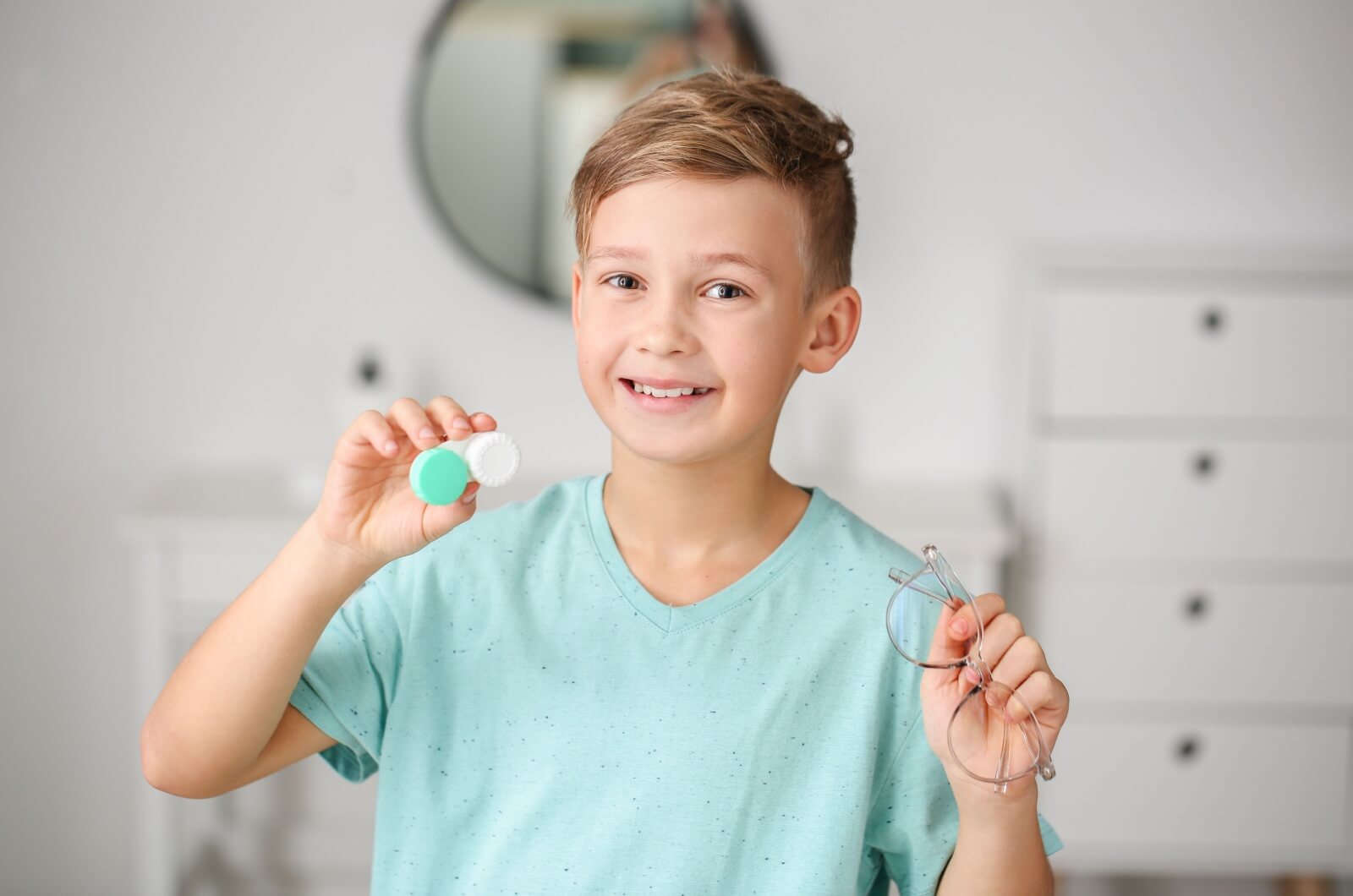 A smiling young child holding a contact lens case in one hand and eyeglasses in the other hand.
