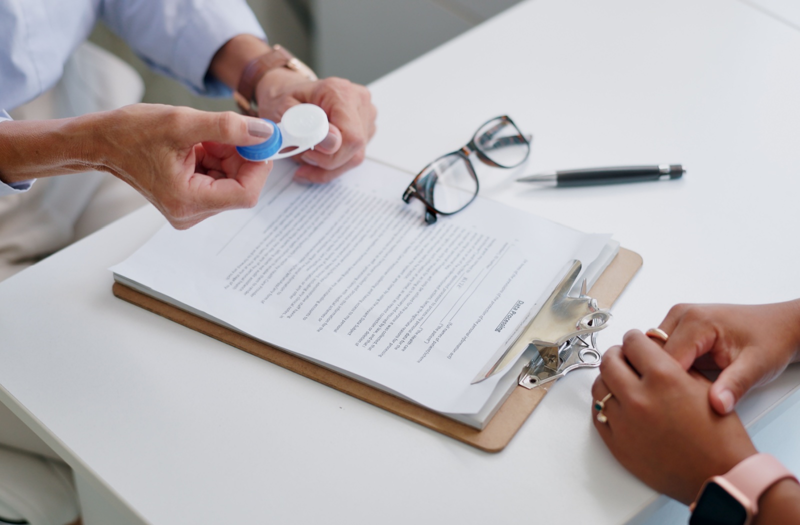 A person holds a contact lens case above a clipboard with glasses and paperwork nearby.