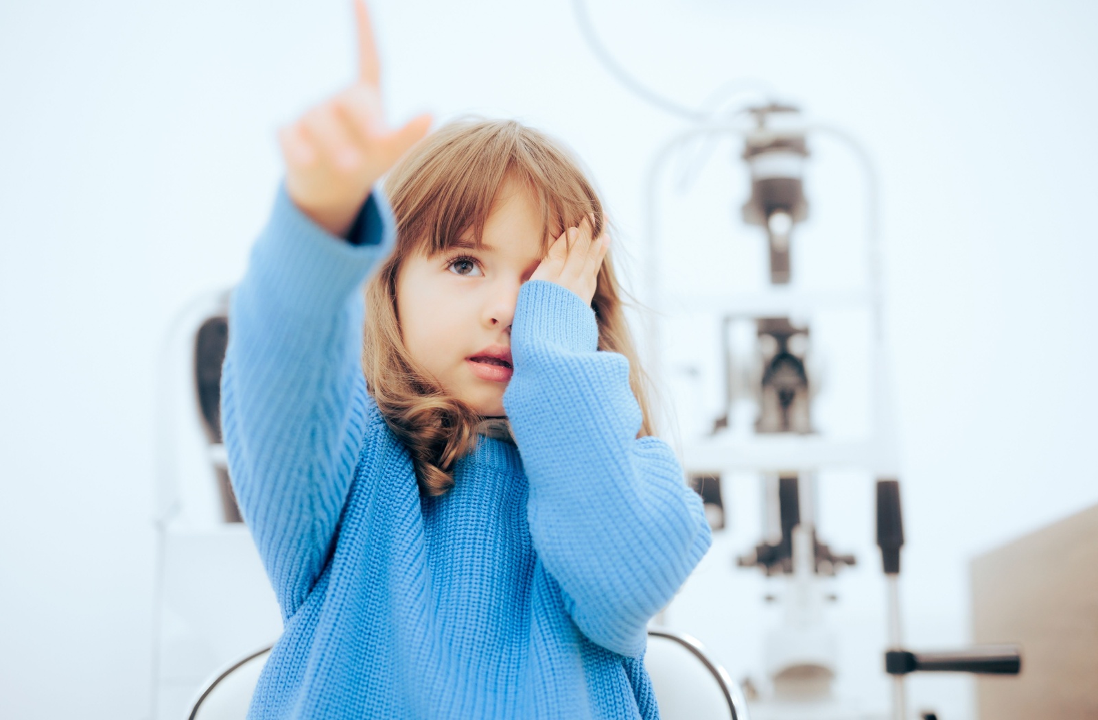 Young girl covering one eye and pointing at a vision chart during an eye exam to test for myopia or astigmatism.