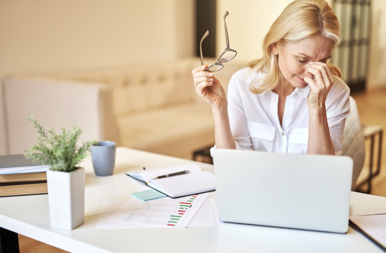 A person sitting at a desk with a laptop and holding their glasses while they rub their dry eyes